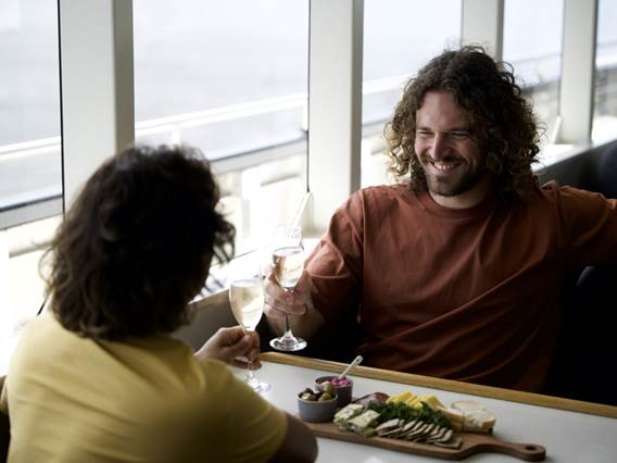A couple cheers champagne over a cheeseboard, while on a cruise boat