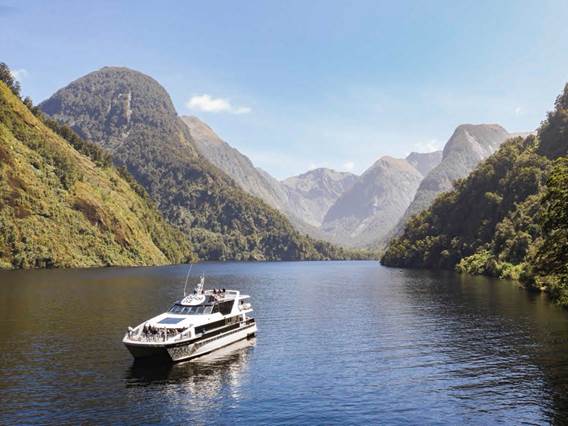 A cruise boat journeys through the water at Doubtful Sound on a clear day, surrounded by mountains 