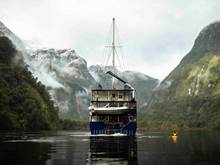 A kayaker paddles next the Fiordland Navigator vessel on Doubtful Sound, surrounded by snowy mountains