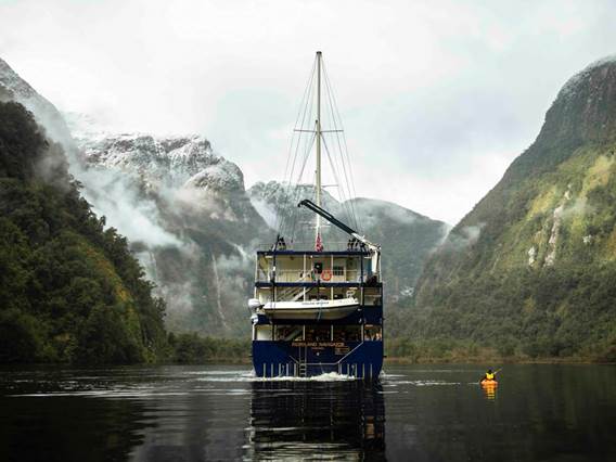 A kayaker paddles next the Fiordland Navigator vessel on Doubtful Sound, surrounded by snowy mountains